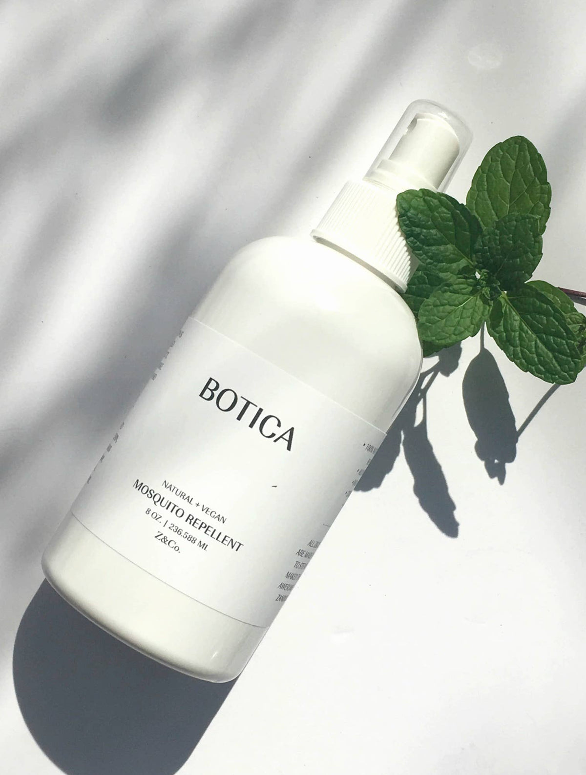 A white bottle of Z&Co. Mosquito Repellent Botica Collection DEET-free spray with mint leaves on its side, casting a shadow on a white surface in bright sunlight.