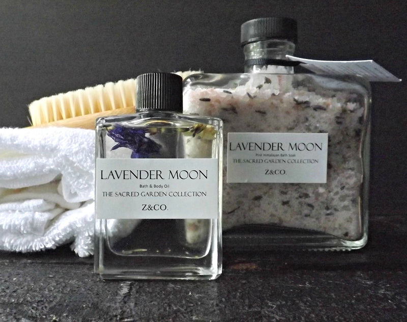 Two Z&Co. bath products on a black background: a clear bottle labeled "lavender moon bath & body" containing essential oils and a jar of "lavender moon the sacred garden collection" Himalayan pink salt.