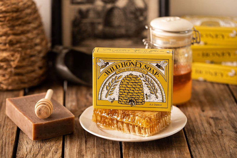 A jar of Primitive House Farm Wild Honey Soap, a propolis-enriched honeycomb slab, and a wooden honey dipper on a table, with a box displaying "Wild Honey" and illustrations of bees and flowers.