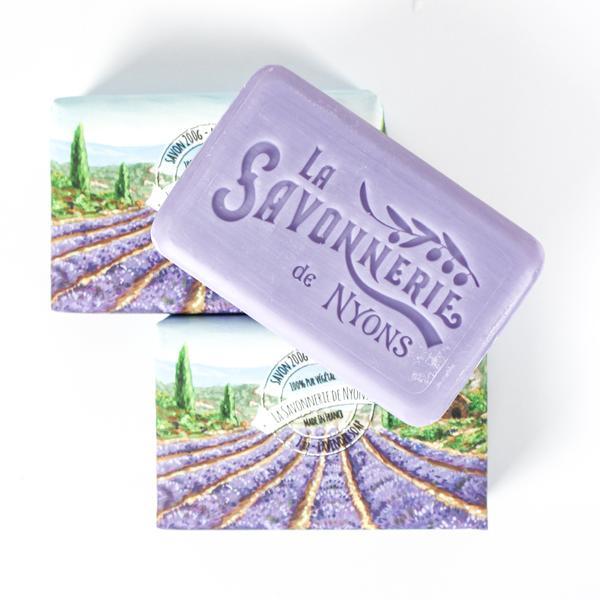 A bar of purple, Lavender scented soap labeled "La Savonnerie de Nyons" rests in front of an illustrated lavender field backdrop, emphasizing its floral essence.