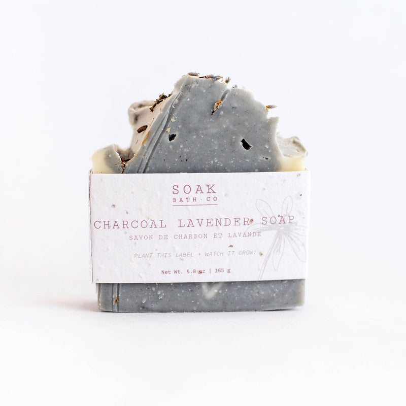 A bar of SOAK Bath Co. - Charcoal Lavender Soap, featuring a textured top with specks, wrapped in labeled white and pink zero waste paper.