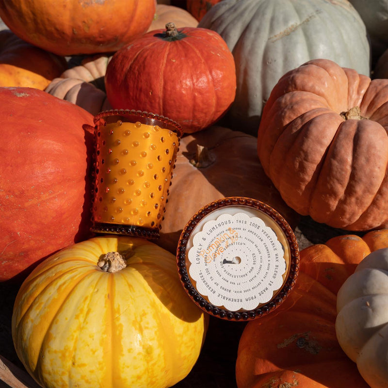 A variety of colorful pumpkins and gourds, including red, orange, and yellow, are displayed with a Simpatico NO. 28 Pumpkin & Clove Hobnail Glass Candle holder and a zodiac-themed plate in a fall setting.