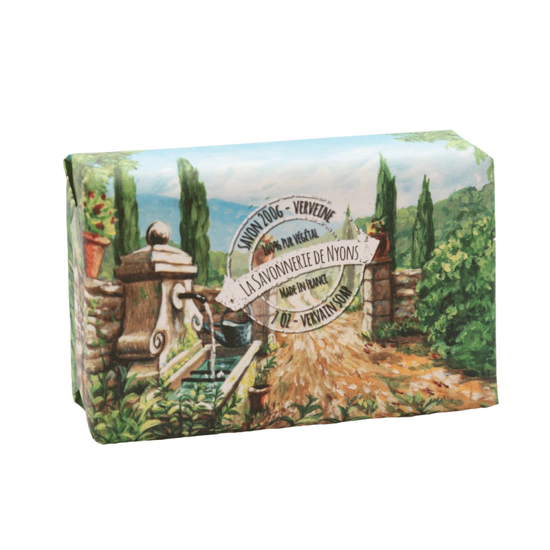 A La Savonnerie de Nyons Verveine Fraiche (Fresh Verbena) 200g soap in paper featuring a painted pastoral scene labeled "la savonnerie de nyons", with illustrations of olive trees, a rustic wooden cart, and Mediterranean landscape elements, now enhanced with organic shea butter.