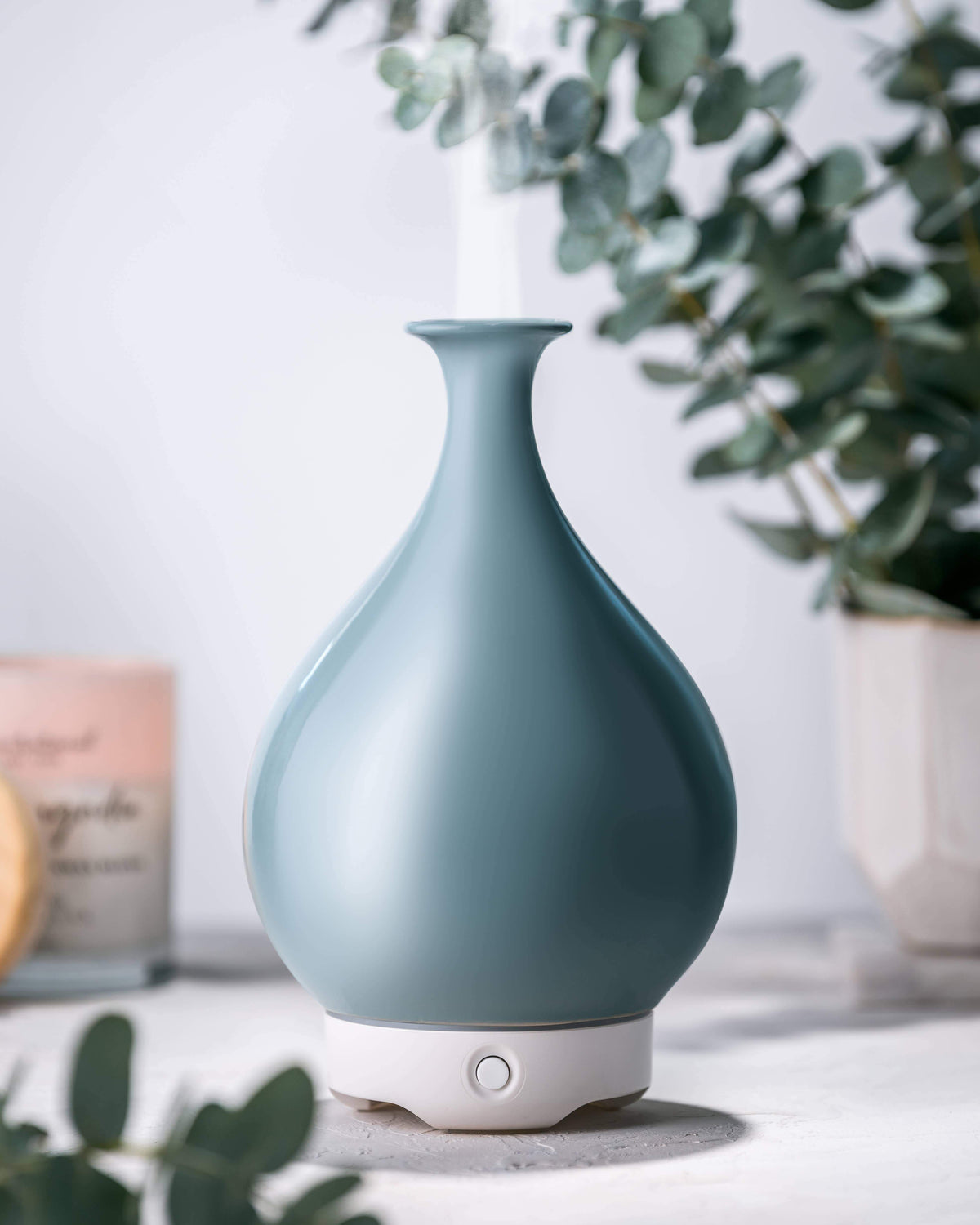 A sleek, soft blue Woolzies Green Glass Vase Diffuser with ultrasonic technology and a teardrop design, positioned on a light tabletop surrounded by eucalyptus leaves with a hint of blurred greenery.