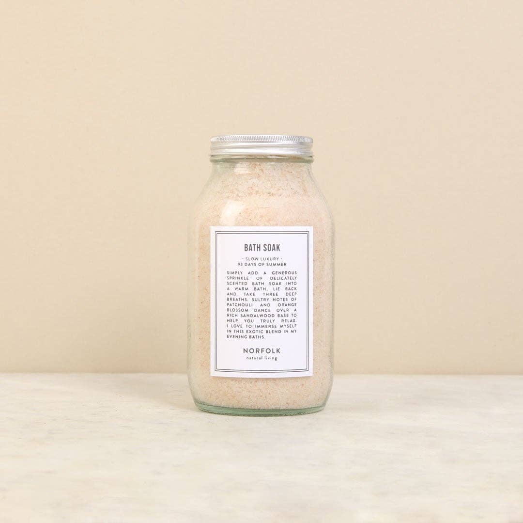 A clear glass jar filled with Norfolk Natural Living Coastal Walks Bath Soak (Himalayan Salt Melody) placed against a pale neutral background. The jar has a simple label with black text detailing the product’s name and ingredients.