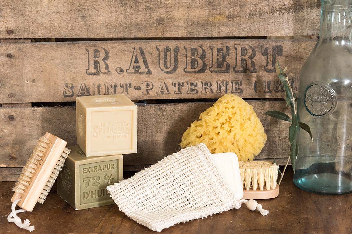 Vintage toiletries on a wooden surface, including natural sponges, La Savonnerie de Nyons cotton soap pocket, a brush, and a loofah, next to an empty glass bottle with a rustic wooden backdrop labeled "La Savonnerie de Nyons".