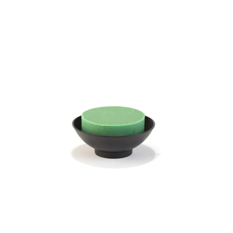 A green round shave soap bar placed in a Three Sisters Apothecary Black Metal Shave Bowl on a white background.