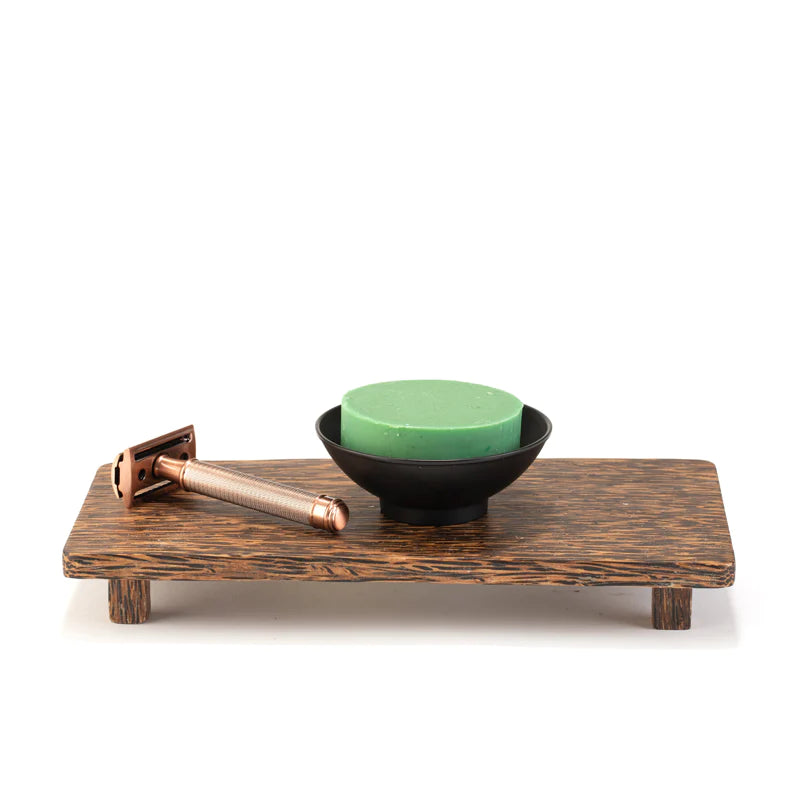 A traditional shaving kit on a wooden tray, featuring a safety razor with a copper handle and a green puck of shave soap in a Three Sisters Apothecary Black Metal Shave Bowl, isolated on white background.