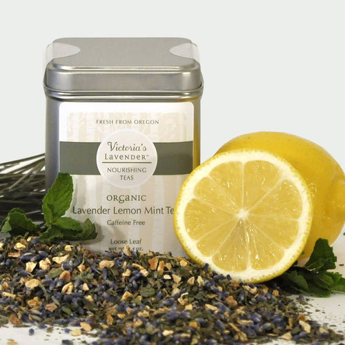 A tin of Victoria's Lavender Organic Herbal Tea - Lavender Lemon-Mint Loose Leaf alongside scattered loose leaf teas, fresh lavender sprigs, and a cut lemon on a white background.