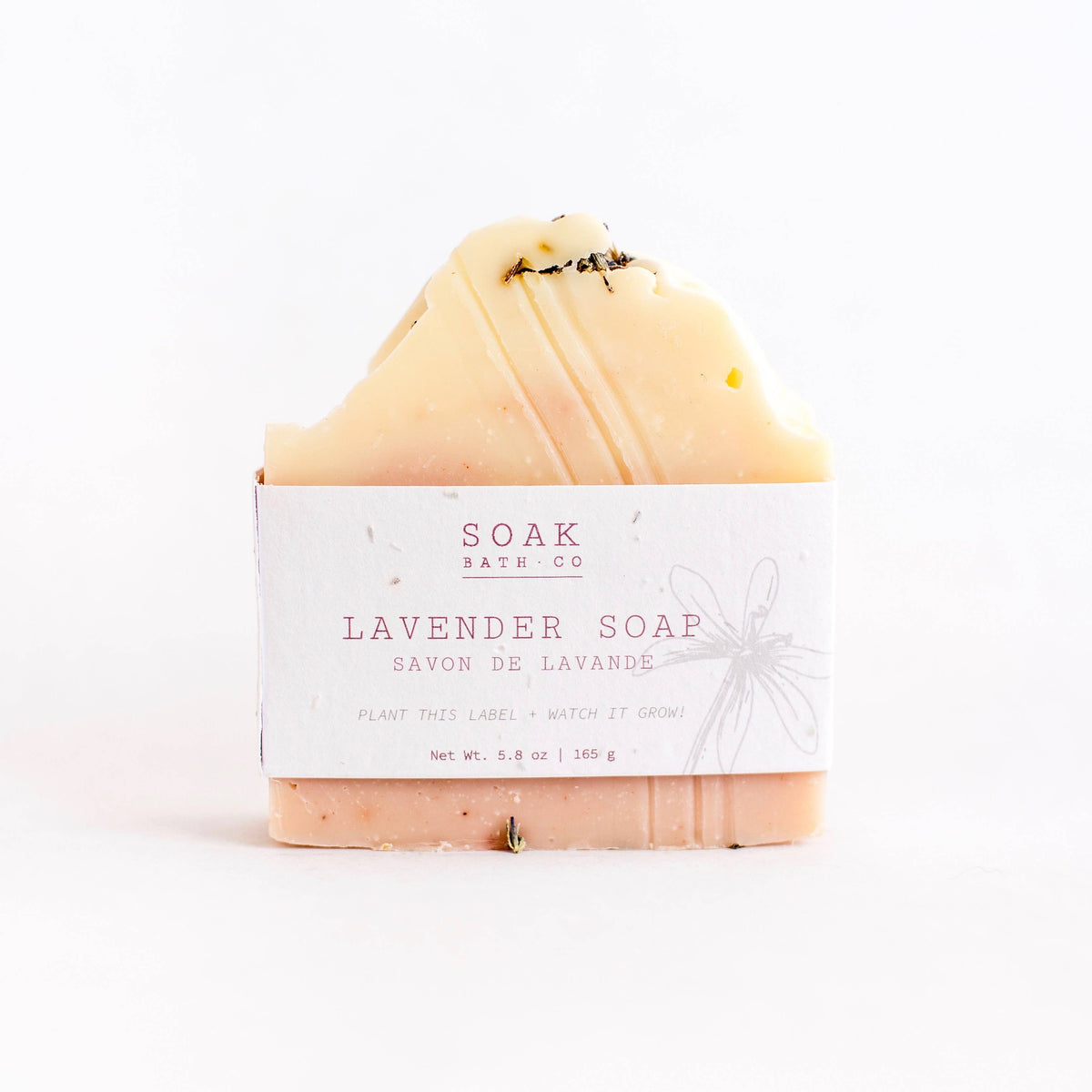 A bar of SOAK Bath Co. - Lavender Soap with a cream and pink gradient, labeled in English and French. There is a bee on the sustainable soap, set against a white background.