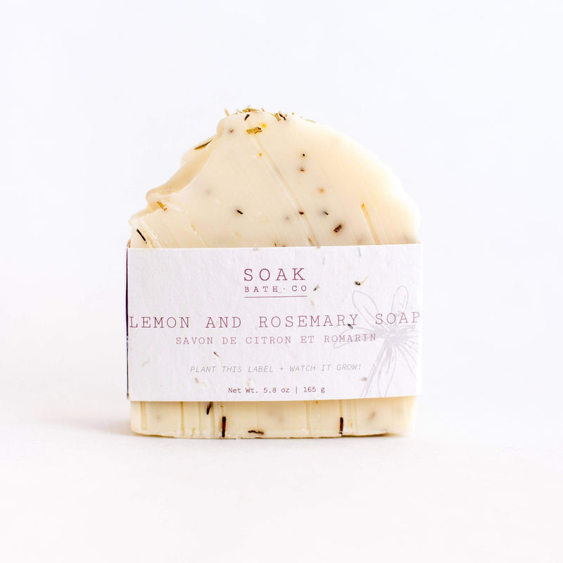 A bar of SOAK Bath Co. - Lemon and Rosemary Soap with a label that reads "soak b.arth." This zero waste soap is textured and sprinkled with herbs, against a white background.