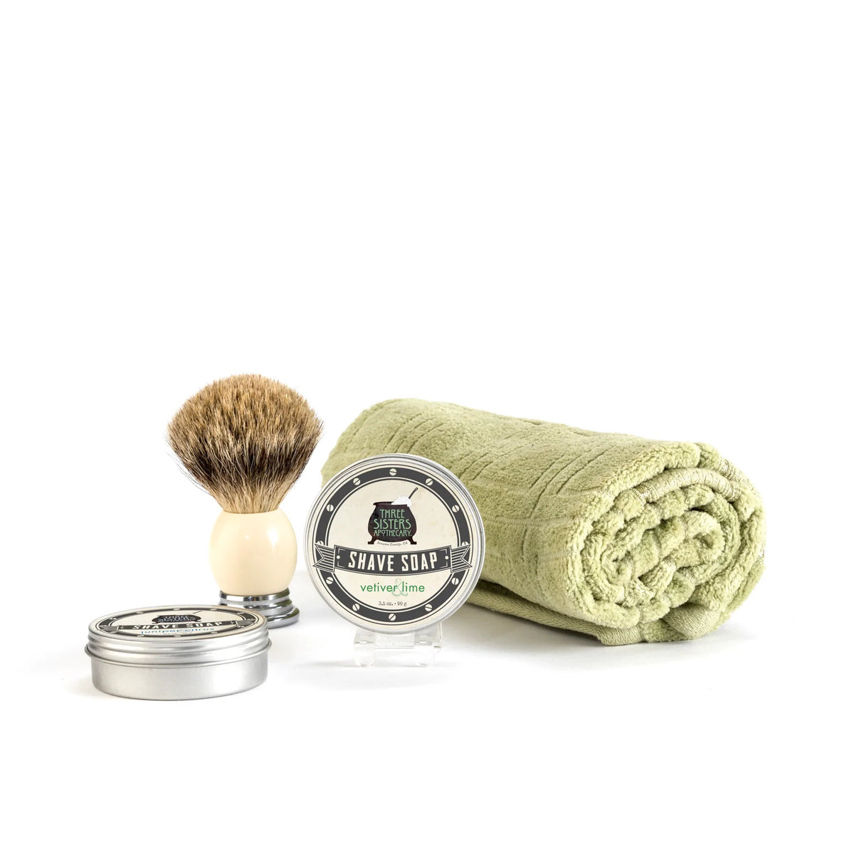 A shaving set displayed on a white background, featuring a badger hair brush, a tin of Three Sisters Apothecary Shave Soap - Vetiver & Lime labeled "austere velvet/lime," and a rolled-up olive green towel.