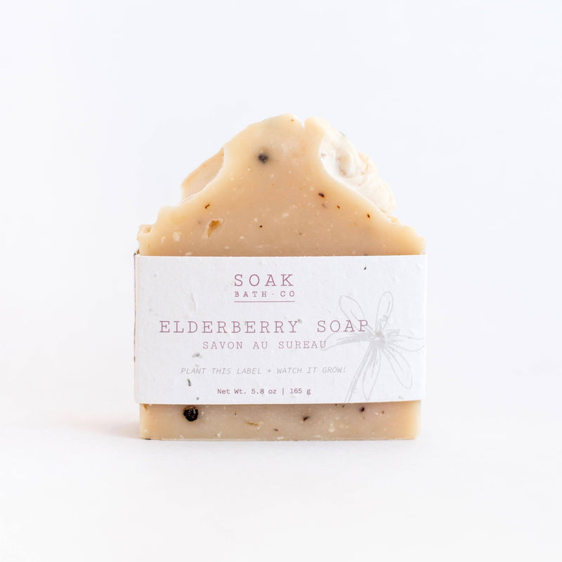 A bar of SOAK Bath Co. - Elderberry Soap with visible specks, wrapped in a sustainable paper label with the brand "SOAK bath co" and the text "plant this label, watch it grow." The background