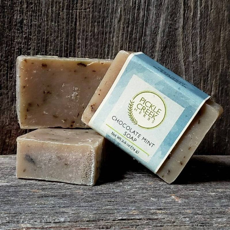 Three bars of Pickle Creek Herbs Chocolate Mint Herbal Soap, stacked against a rustic wooden background. The soaps are natural-toned with visible specks.