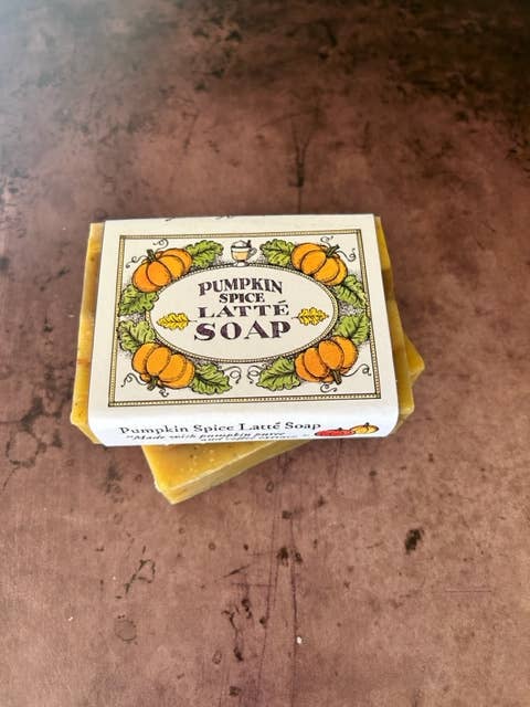 A bar of Primitive House Farm Pumpkin Spice Latte soap infused with coffee extract rests on a weathered surface, featuring a label with orange pumpkins and decorative greenery.