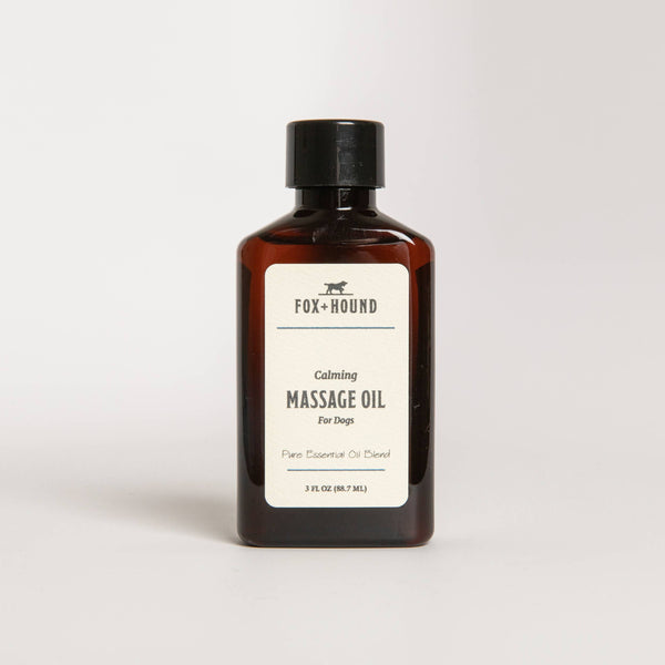 A brown glass bottle labeled "Fox + Hound - Calming Massage Oil For Dogs 3 oz" featuring therapeutic essential oils, in a simple, clean design on a white background.