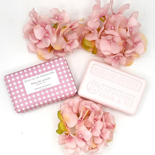 Two bars of Senteurs De France Vichy Cherry Blossom Soap labeled in French, one with its matching pink and white checked packaging, surrounded by delicate cherry blossom flowers on a white background.