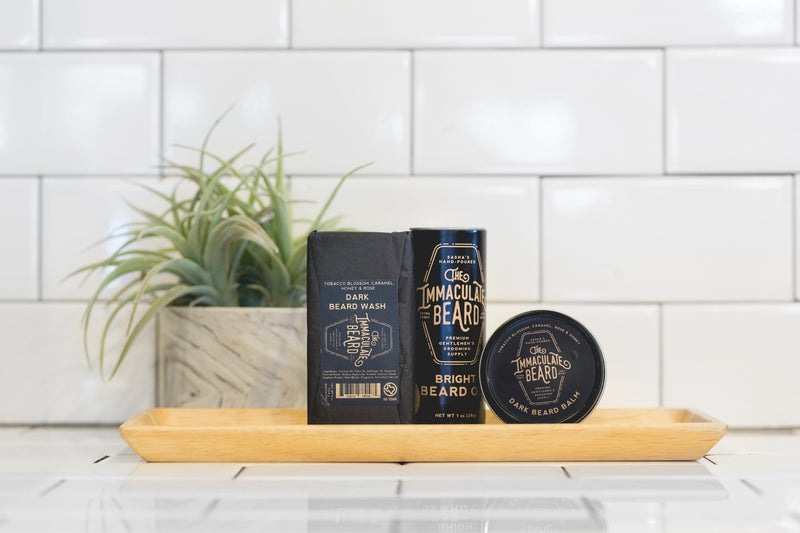 A neatly arranged men's grooming set placed on a wooden tray against a white tiled wall. It includes The Immaculate Beard - Beard Wash Bar with activated charcoal, beard oil, and beard balm from The Immaculate Beard, each labeled in stylish black packaging.