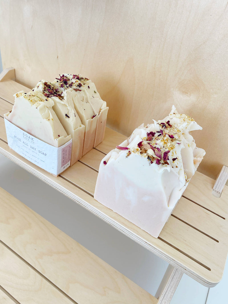 Handcrafted SOAK Bath Co. - Rosé All Day Soap bars with a creamy texture and sprinkled with dried petals, displayed on a wooden shelf; each bar is wrapped in zero-waste, sustainable packaging showing brand details.