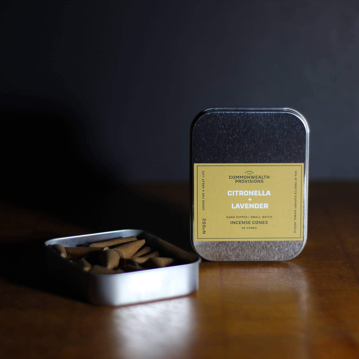 A tin container labeled "Commonwealth Provisions Citronella + Lavender Incense Cones" with the lid open, showing the cones inside, placed on a wooden surface against a dark background.