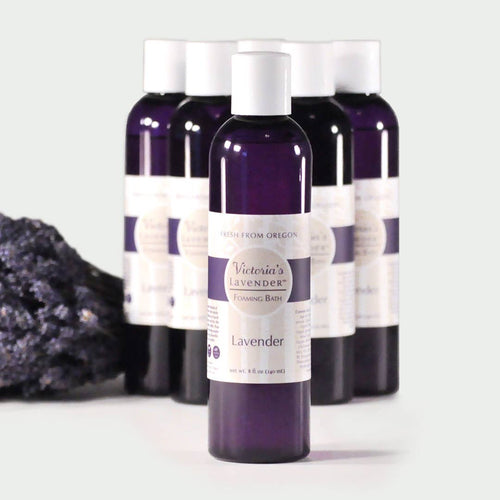 Five bottles of Victoria's Lavender - Lavender Foaming Bath, enhanced with essential oils, are arranged in a line on a light gray background, with a focus on the front bottle displaying a lavender label.