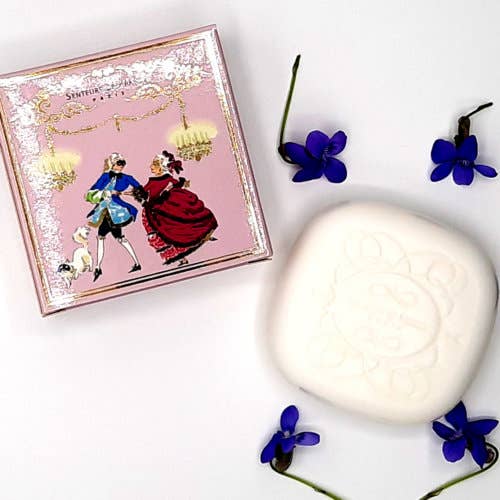 A decorative Senteurs De France Violet Soap "Masked Ball" with an embossed flower design, positioned near a book with a vintage illustration of a dancing couple on the cover, surrounded by scattered purple flowers.