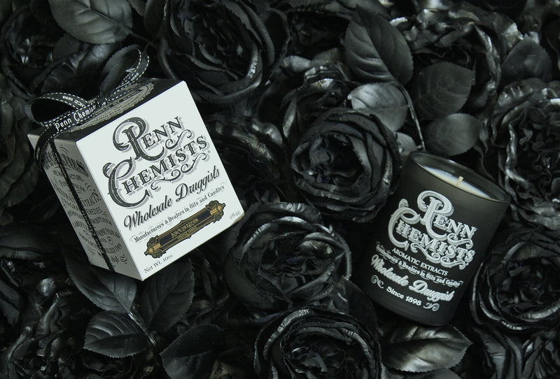 A moody, monochrome image of a Penn Chemists Classic Candle - Black Gardenia box and container placed amid a bed of black silk roses, exuding an elegant, gothic aesthetic with hints of Tahitian Gardenia.