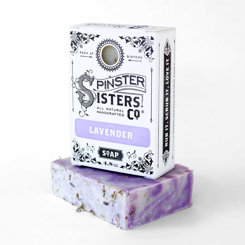 A bar of Spinster Sisters Bath Soap - Lavender with Lavender Blossoms topped with dried lavender pieces, placed next to its packaging. The black and white box features ornate designs and the text "Spinster Sisters Co. All Natural Handcrafted.