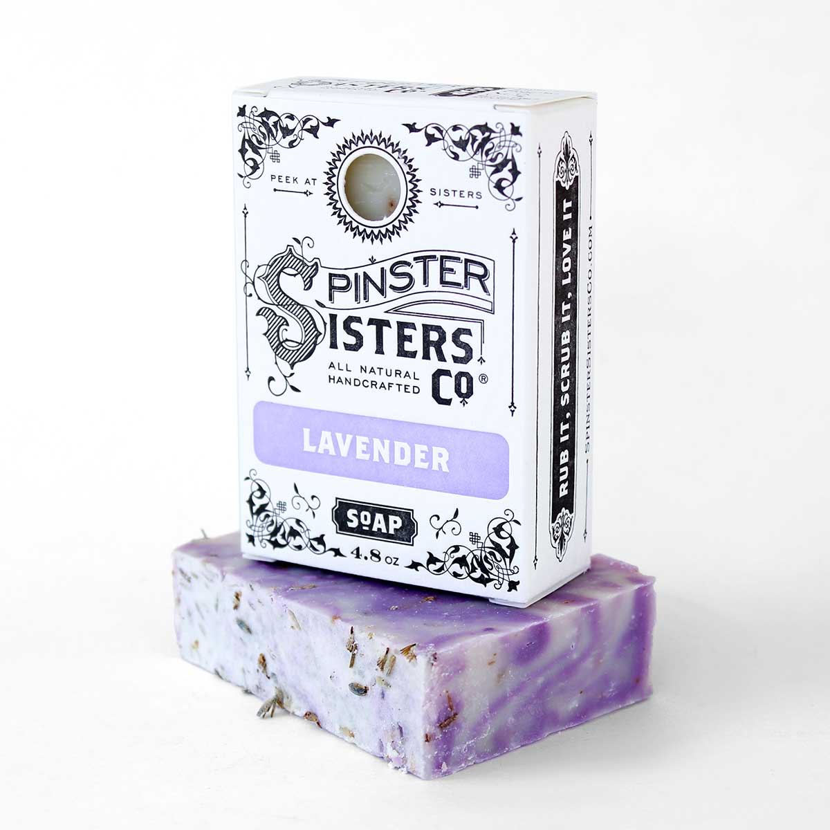 A bar of Spinster Sisters Bath Soap - Lavender with Lavender Blossoms topped with dried lavender pieces, placed next to its packaging. The black and white box features ornate designs and the text "Spinster Sisters Co. All Natural Handcrafted.