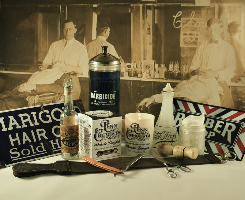 Vintage barber shop display featuring barber tools, bottles of cologne scent, a marigold hair oil sign, and an old photo of barbers in the background, all arranged on a table with Penn Chemists Classic Candle - Cut & Shave from Penn Chemists.