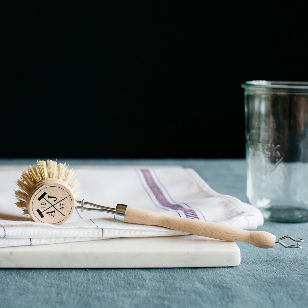 A Andrée Jardin Tradition Handled Dish Brush with natural bristles, made in France, sits on a folded linen towel next to an empty glass jar, all positioned against a dark background.