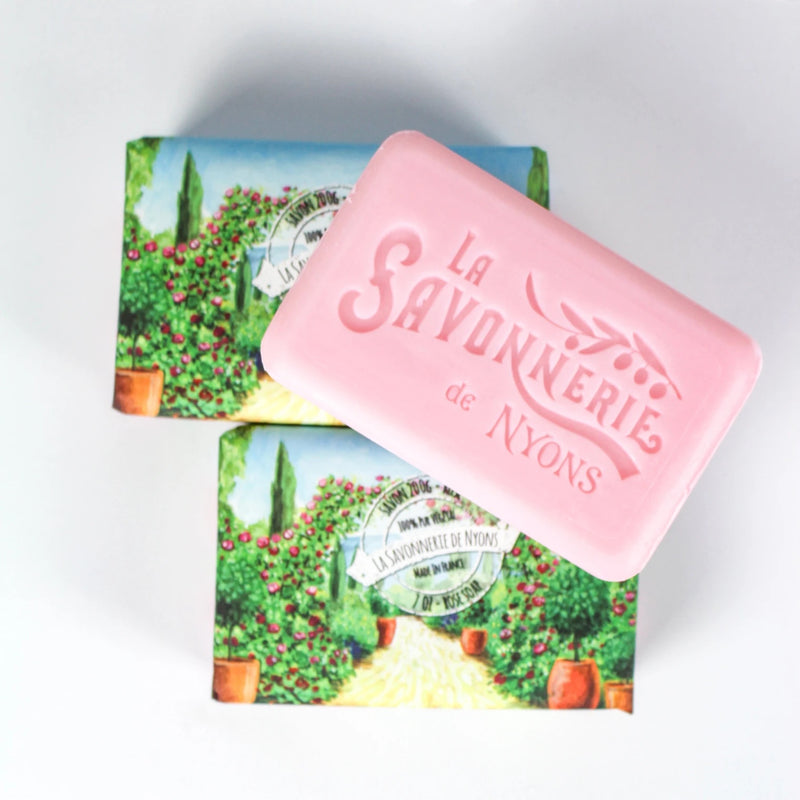 A pink bar of rose scented soap labeled "La Savonnerie de Nyons Jardin de Roses" in front of its packaging, which features an illustrated vineyard scene.