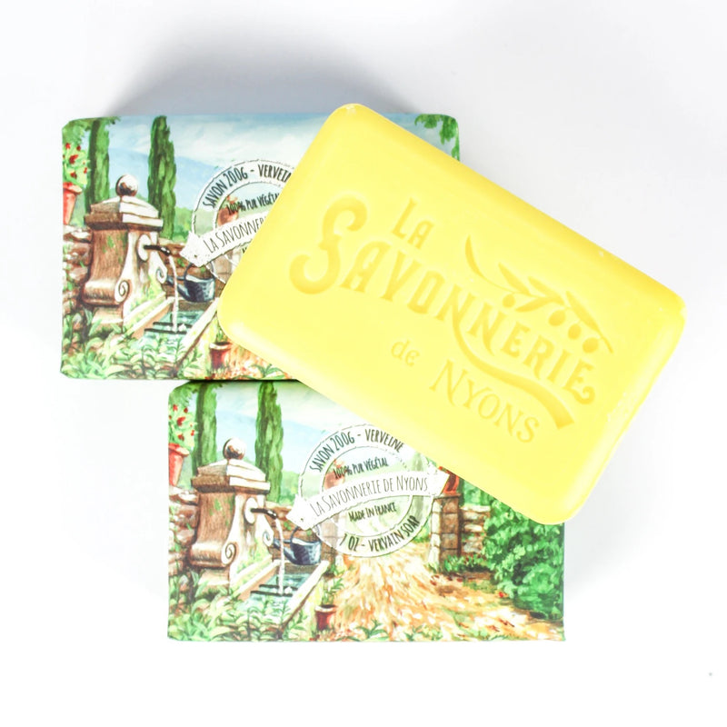 A yellow bar of La Savonnerie de Nyons Verveine Fraiche scented soap labeled "la savonnerie de nyons" resting on two colorful illustrated soap boxes featuring pastoral French scenes.