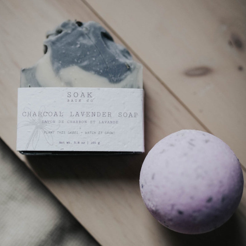 A charcoal lavender soap bar in a paper wrapper next to a SOAK Bath Co. Lavender Bath Bomb on a wooden surface, with the soap label featuring plantable instructions.
