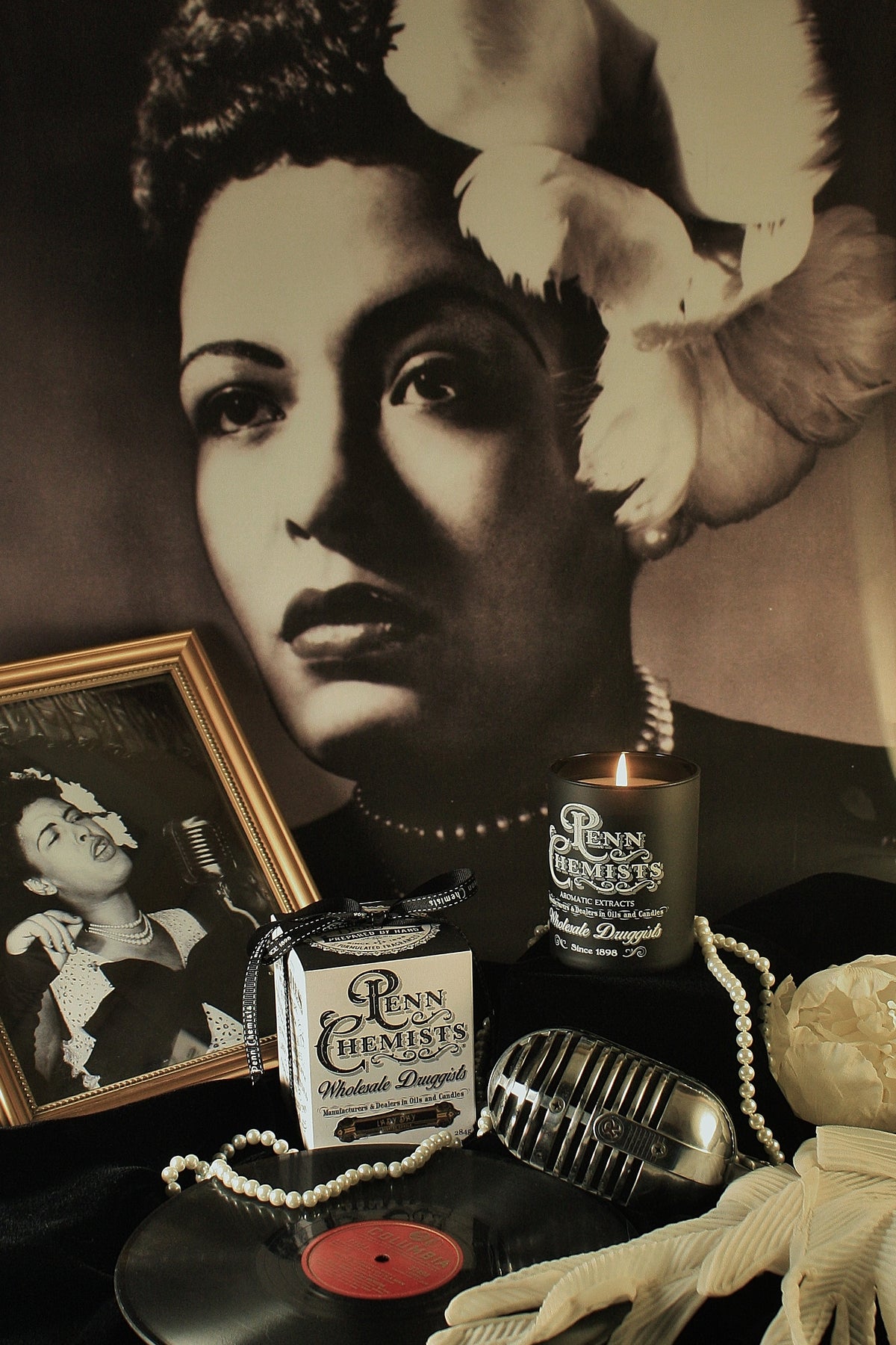 Vintage tribute setup featuring a large portrait of a classic film actress, surrounded by old-time accessories including a microphone, vinyl record, pearls, memorabilia, and a Penn Chemists Classic Candle - Lady Day.