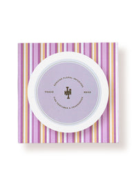 A round floral-infused Lollia Relax body butter box with a lavender and white color scheme, displaying Margot Elena's trademark logo, resting on a purple and white striped background.