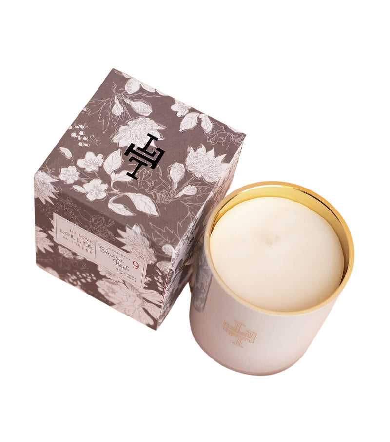 A Lollia In Love No. 09 Perfumed Luminary Candle in a handblown glass holder with embossed initials next to its floral-patterned packaging box, isolated on a white background.品牌：Margot Elena