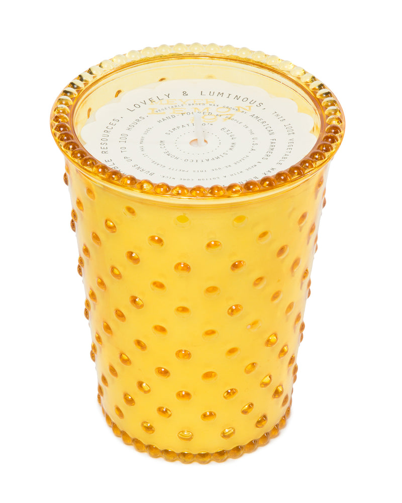 A vibrant yellow, hobnail glass candle with a white label on the lid, isolated on a white background. This Simpatico NO. 97 Meyer Lemon Hobnail Glass Candle is infused with a citrus fragrance.