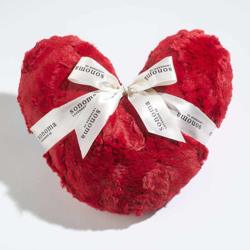 A plush red Sonoma Lavender Comforting Heart Pillow tied with a white ribbon, printed with the word "somnium" on it, and infused with a calming lavender scent, against a white background.