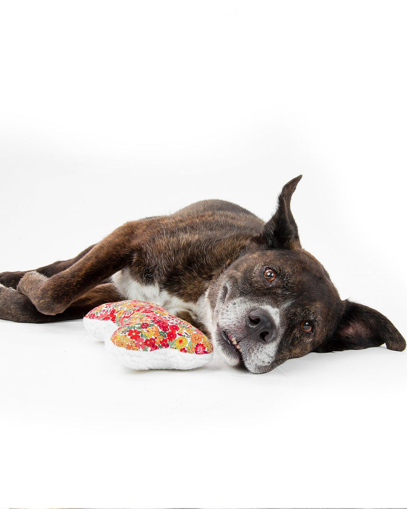 A brindle-coated dog lying on its side on a white background, head resting on a MODERNBEAST Lavender Zenbone - Red Floral stuffed toy, looking directly at the camera with a relaxed expression.