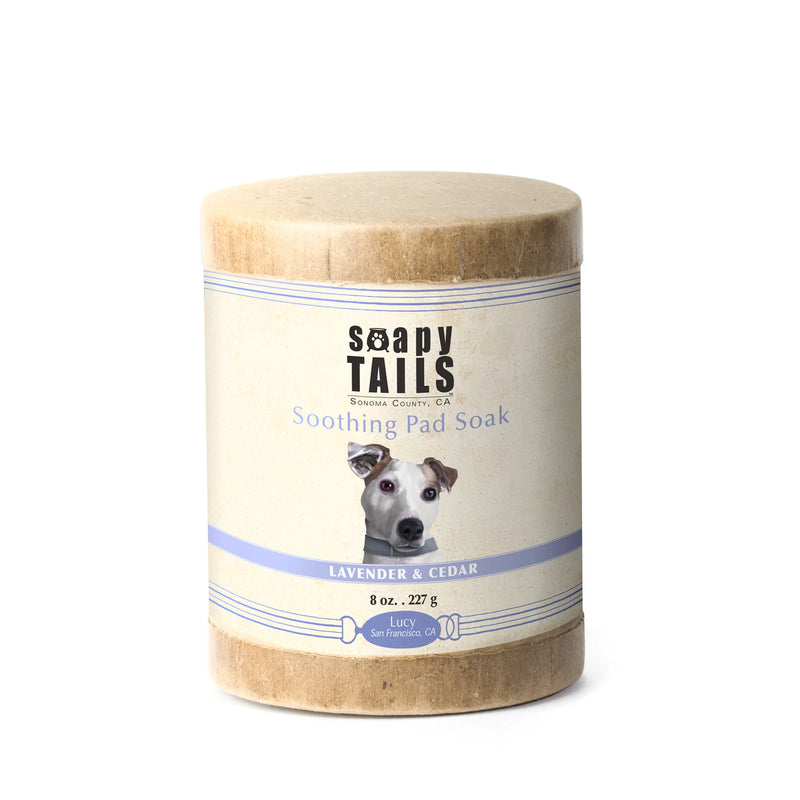 A cylindrical container of "Three Sisters Aphothecary Soapy Tails Dog Pad Soak" for dogs, featuring lavender and cedar scents. The label includes an image of a dog's face and product details on it.