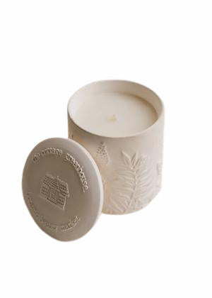 A beige soy wax blend candle with a decorative lid, featuring embossed tropical leaf patterns and textual branding from The Cottage Greenhouse Himalayan Salt & Rosewood Ceramic Candle by Margot Elena, isolated on a white background.