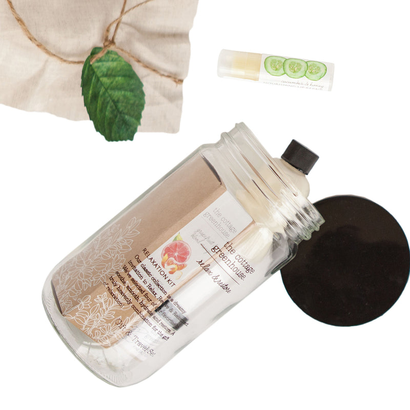 A transparent cosmetic bottle labeled "Margot Elena Cottage Greenhouse Fruit Relaxation Kit" with a green leaf and a rolled-up label next to two small lotion tubes on a white background.