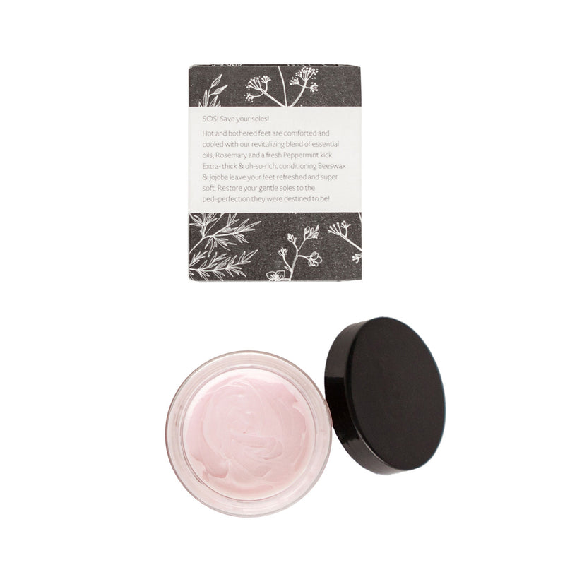 Top-down view of an open jar of The Cottage Greenhouse Rosemary Mint Rescue Foot Cream next to its lid and a box with botanical illustrations and product details, including Peppermint essential oil, displayed on a white background by Margot Elena.