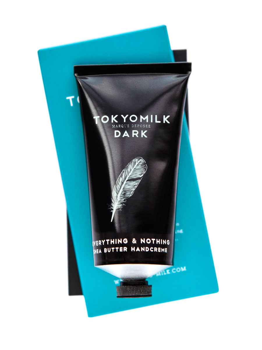 A tube of Margot Elena's TokyoMilk Dark Everything & Nothing Handcreme shea butter hand cream resting on a teal book, against a white background. The tube is black with a white feather design.
