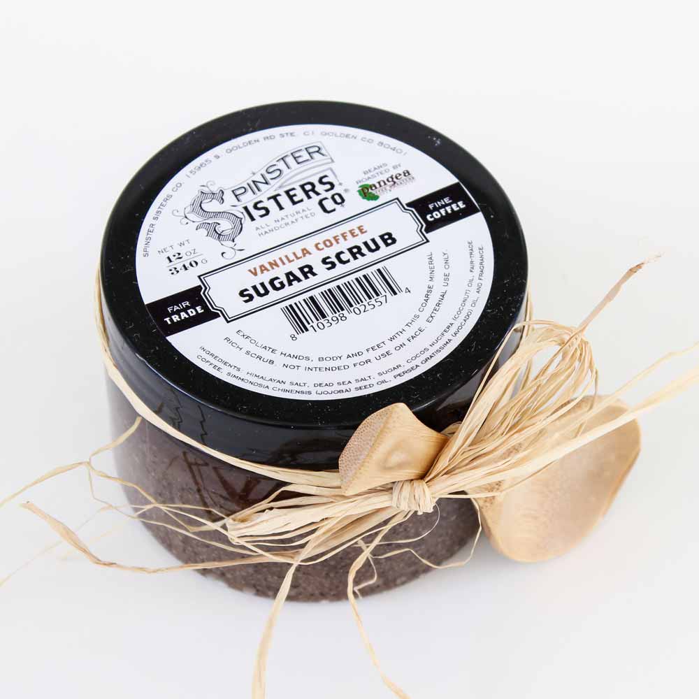 A jar of Spinster Sisters, Co. Vanilla Coffee Sugar Scrub designed to exfoliate skin, with a golden spoon tied to it with a raffia ribbon, against a white background.