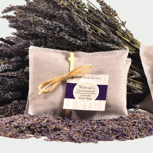 A set of 3 Victoria's Lavender Dryer Sachets tied with a yellow ribbon and a label reading "Victoria's lavender dryer sachets, fresh from Oregon," surrounded by dried lavender flowers, perfect for the laundry room.