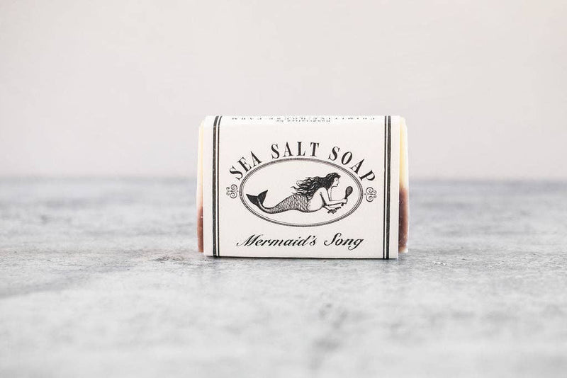 A bar of sea salt soap labeled "Forty Fathoms Mermaid Soap" from Primitive House Farm, infused with Brazilian purple clay, sits on a plain surface. The packaging features an illustration of a mermaid in a circular frame.