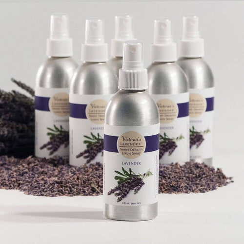 A collection of silver bottles labeled "Victoria's Lavender - Sweet Dreams Lavender Linen Spray," enriched with lavender essential oil, arranged on a surface, with dried lavender bundles in the background.