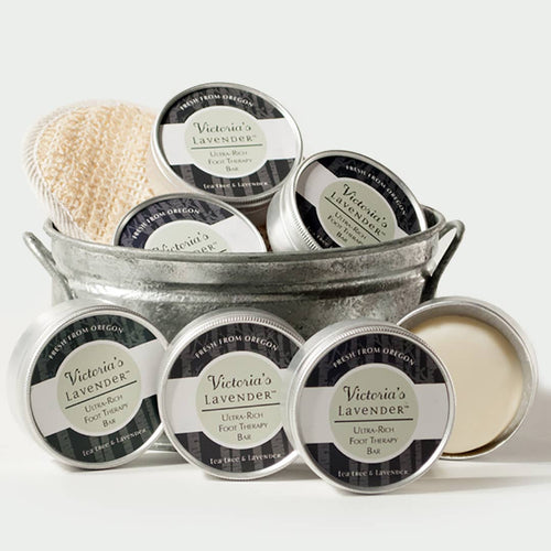 A collection of Victoria's Lavender lavender foot therapy lotion bars, infused with tea tree oil, and a scrub brush, neatly arranged in a rustic metal bucket on a white background.